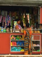 A small shop/kiosk with colourful products displayed on shelves, and a person wearing a traditional sari is standing at the counter.